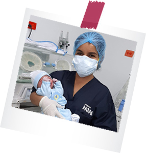A health worker in navy blue Project Hope scrubs, a face mask, gloves, and cap smiles as they hold a newborn baby in a blue outfit and knit hat